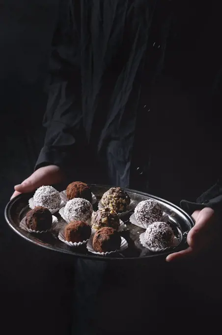 Variety of homemade dark chocolate truffles with cocoa powder, coconut, walnuts on vintage tray in kid's hands in black shirt. Dark background.