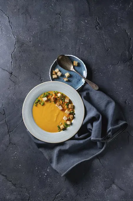 Plate of vegetarian pumpkin carrot soup served with croutons and onion on textile napkin over dark texture background. Top view with space