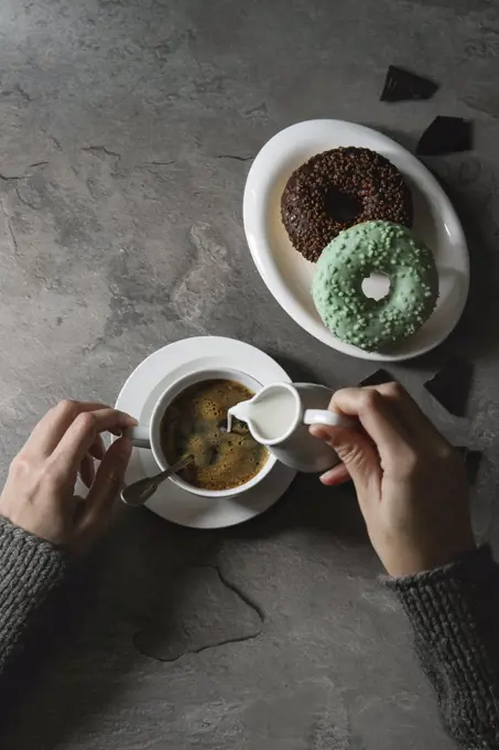 Plate of colorful glazed donuts with chopped chocolate, cup of black coffee, jug of milk over gray texture table. Female hands pouring cream. Flat lay with space. Rustic style
