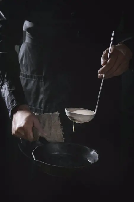 Man chef in black apron pouring dough from ladle for cooking pancakes in cast-iron pan. Dark rustic style.