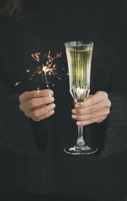 Christmas or New Year celebration concept. Woman in dark grey woolen sweater holding glass of champagne and traditional festive sparklers in hands, close-up