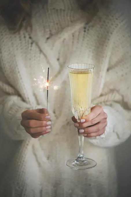 Christmas or New Year celebration concept. Woman in white woolen sweater holding glass of champagne and sparkler in hands, close-up