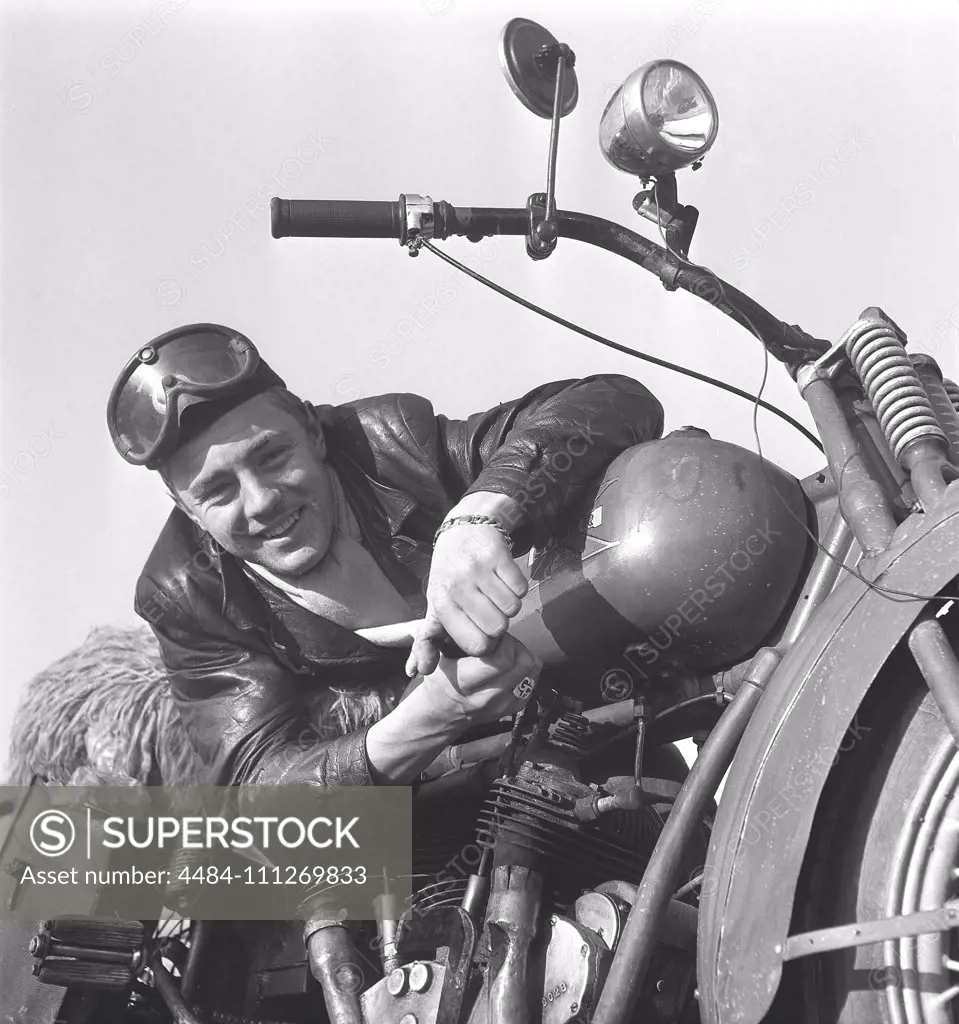 Motorcyclist in the 1950s. A young man wearing glasses and a leather cap by his motorcycle. The motorcycle is made by the american company Excelsior in Chicage and the model is Super X 1925, and was americas first motorcycle with a v-twin engine. Sweden 1950s ref 10-09-11