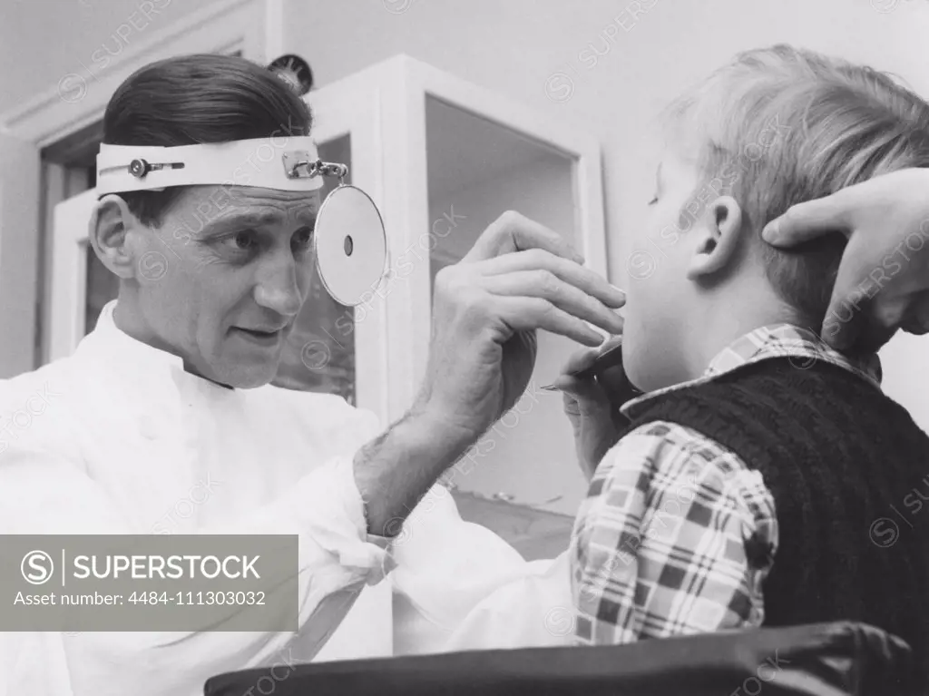 Health care in the 1950s. A doctor is examining a young boy and looks down his throat to see if there is an infection. A special reflecting mirror in his forehead focus the light and makes it easier to see. Sweden 1950s