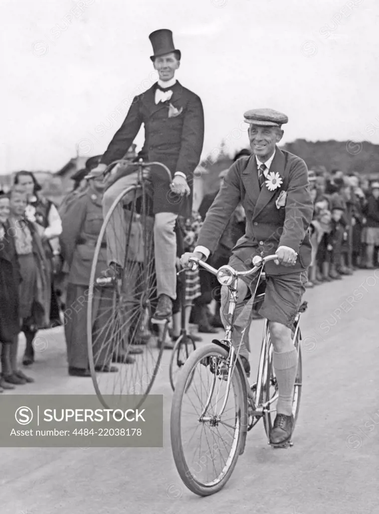 Penny farthing bicycle. A man is riding a penny-farthing bicycle besides a man on a regular bicycle. 1930s