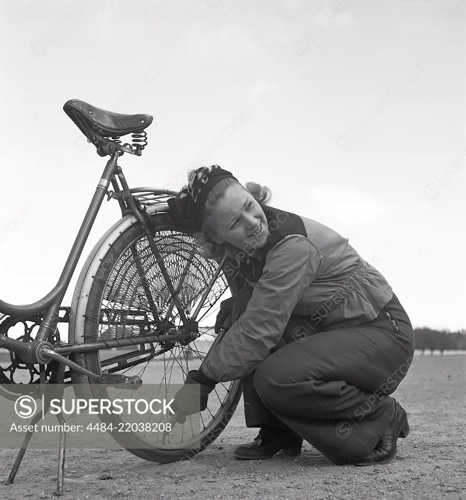 1940s couple with a bicycle. A young woman is pumping air into the back tire of her bicycle. She is wearing the typical sports clothing of the 1940s with long trousers and a short jacket. Notice the protection net mounted over the back wheel to prevent hanging clothes from getting caught in the wheel. Sweden 1942. Photo KristofferssonA89-1