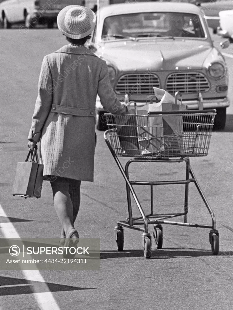 Shopping in the 1970s. A lady is leaving the supermarket and heading for her car pushing the shopping cart on the parking lot. Sweden 1970
