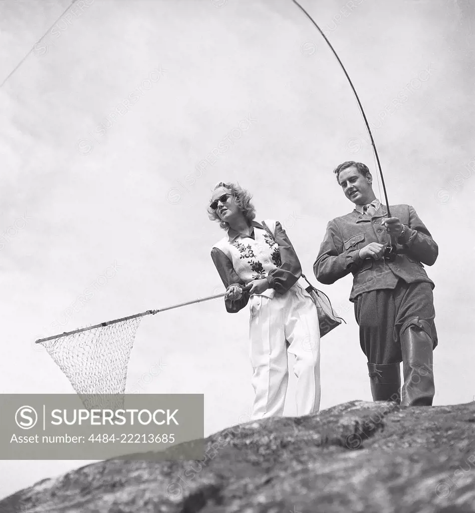 Fishing in the 1950s. A man with a fishing rod looks as if he has a fish on  the hook, with his girlfriend ready with a fishnet. Photo Kristoffersson  ref BF76-4 Sweden