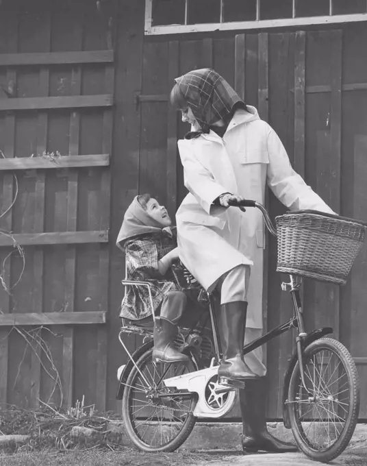 Rain in the 1960s. A young smiling woman is dressed in rain clothes and rides a bicycle with her daughter. Sweden 1967