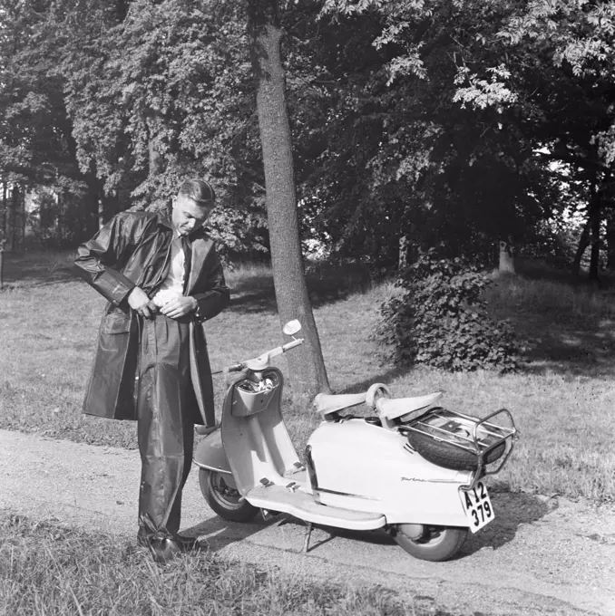 Rain in the 1950s. A young man prepares for a tour on his scooter motorcycle NSU Prima. He dresses in a raincoat and waterproof trousers. Sweden September 1959