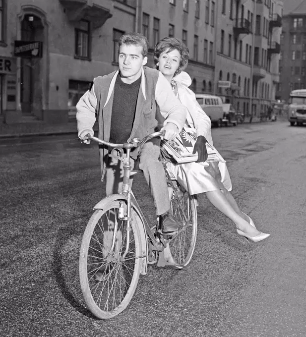 1960s cyclist. A young man on his bicycle has a fashionable dressed woman sitting on the rack on the rear wheel. She is the swedish singer Lill-Babs Svensson. Sweden 1961. Photo Kristoffersson CP83-5 