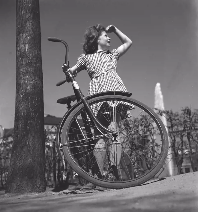 1940s girl with bicycle. A teenage girl, dressed in a chequered summer dress, is standing by her bicycle a sunny day. Sweden May 1940. Photo Kristoffersson 133-2