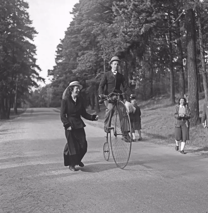 Penny farthing bicycle. A man is riding a penny-farthing bicycle and a woman is running by his side as if she is afraid he will fall. Sweden 1940s Photo Kristoffersson K131-2
