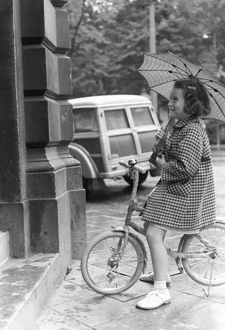 1950s girl on a bicycle. A girl is trying out a new bicycle outside the bicycle shop. She sits happily on the bicycle in the rain under her umbrella. Sweden 1954. Photo Kristoffersson 2A-29