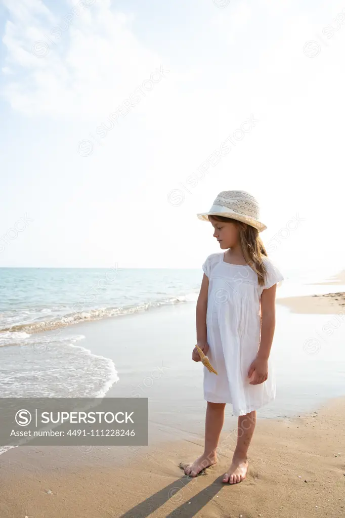 Pretty serious girl with long hair in hat holding starfish on seashore in summer day
