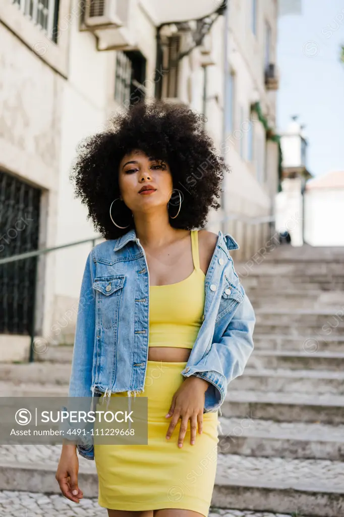 Beautiful African American woman in yellow suit standing on stairs and looking at camera on urban background