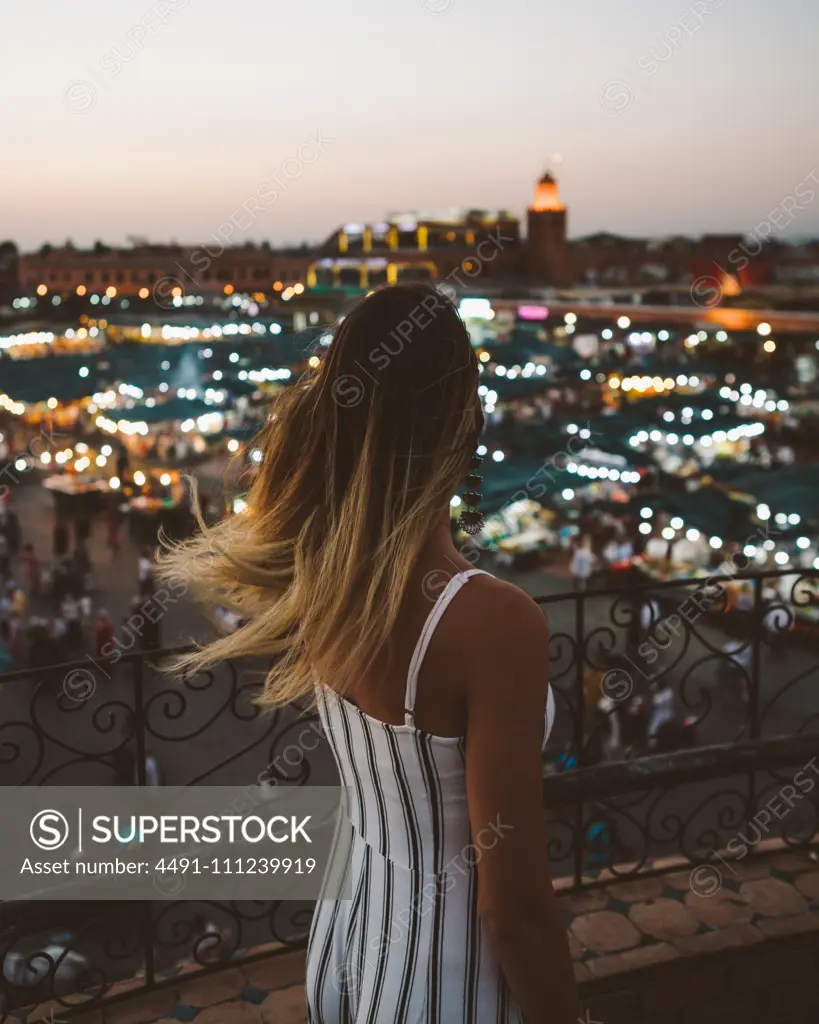 Back view of trendy woman with flying hair standing on balcony against bright lights of Moroccan city in twilight