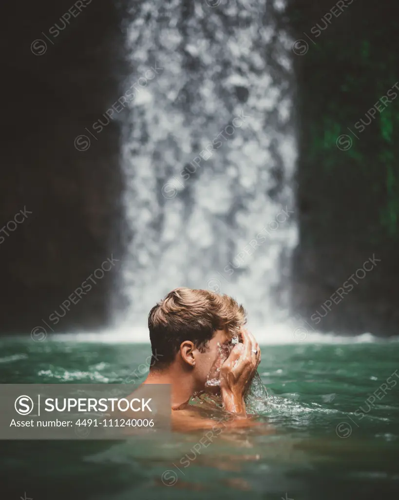 Side view of man washing face while swimming in clear water of lake with waterfall on background, Bali