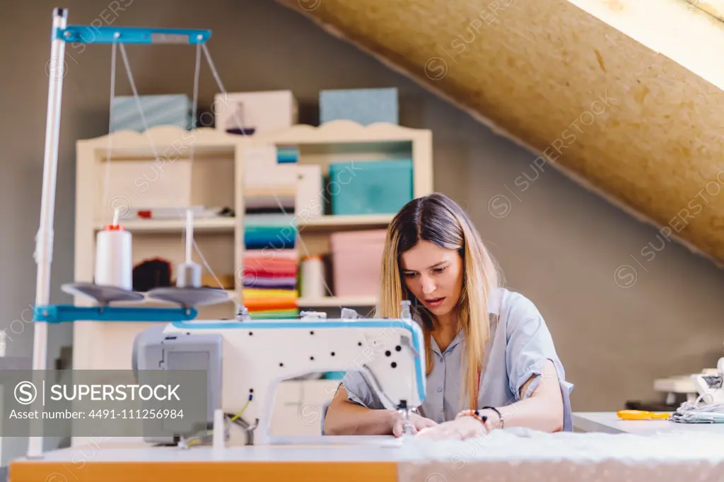 Concentrated female tailor sewing on machine while sitting at table on blurred background of workshop