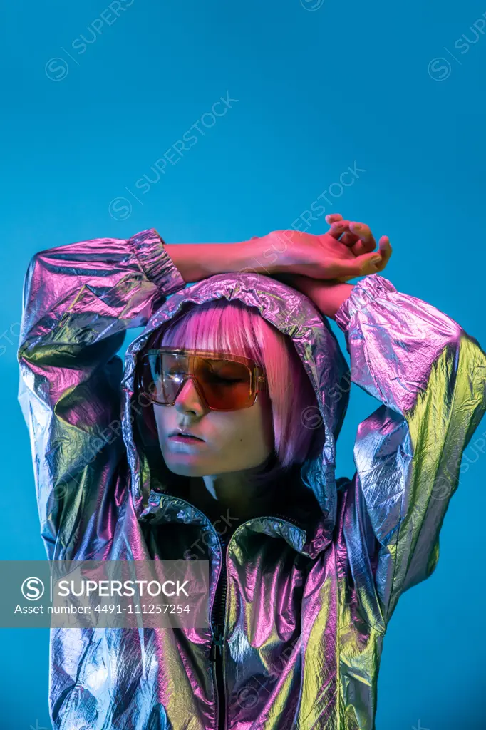 Trendy young Japanese woman with purple hair standing in sparkly silver jacket and red sunglasses on blue background