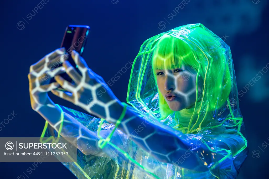 Young Asian woman in futuristic wear and green wig taking selfie on smartphone in fluorescent light