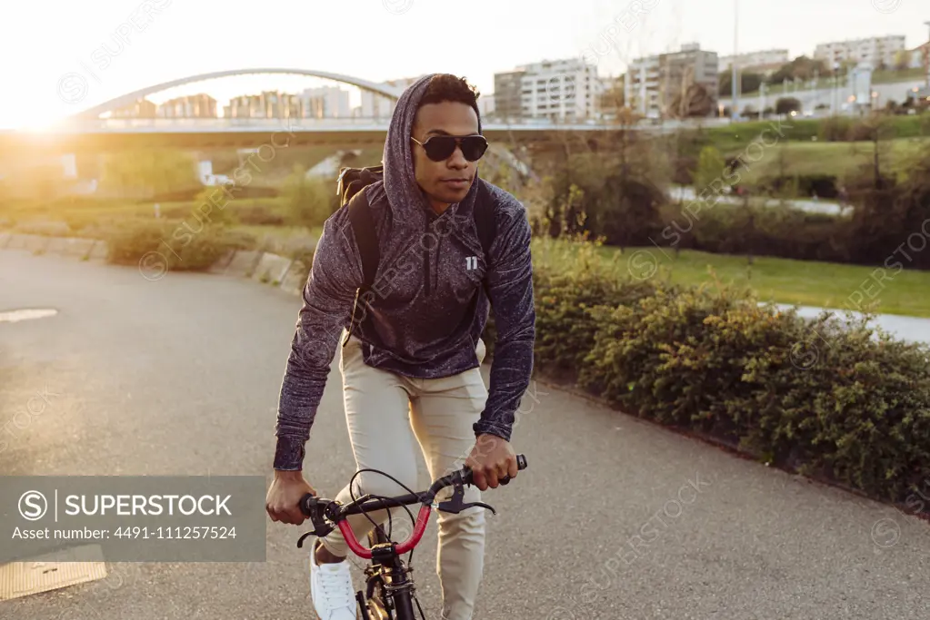 Young African American handsome man in casual outfit and stylish sunglasses riding bicycle near bridge in bright back lit
