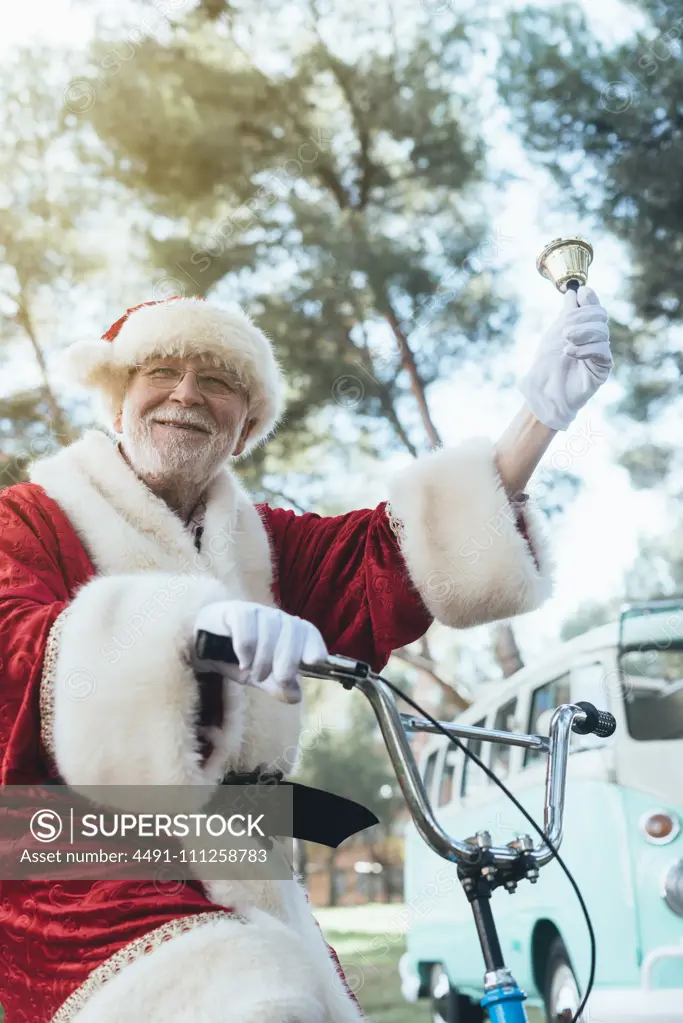 From below senior man in costume of Santa Claus sitting on cycle, ringing bell and looking at camera