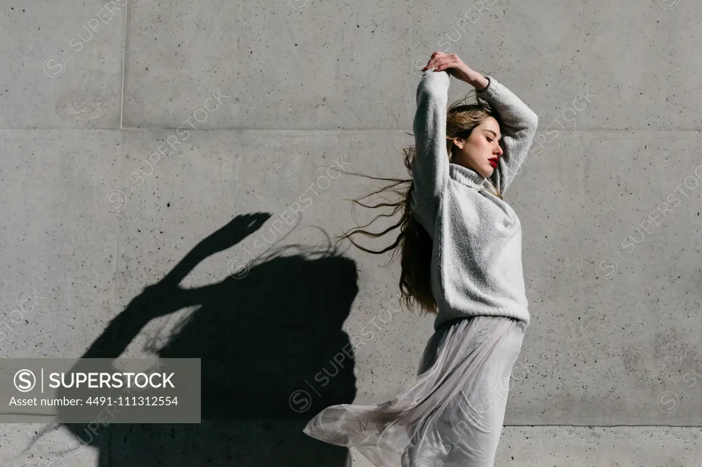 Side view of young female model in stylish warm sweater and skirt closing eyes raising elbow while standing against gray wall on city street 