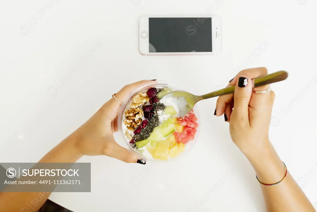 From above anonymous female enjoying healthy dish with fruits and walnuts over table with smartphone in restaurant