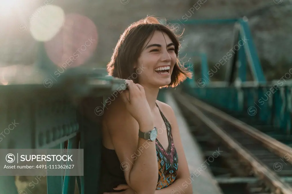 Cheerful young woman looking away with laugh holding iron fence handrail in sunny day on railway bridge