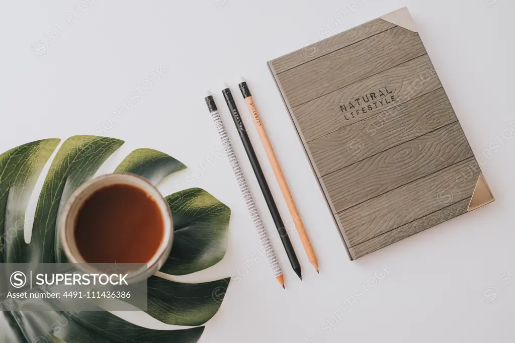 From above stylish notebook with wooden cover composed with row of pencils and mug of hot drink on green Monstera leaf on white background
