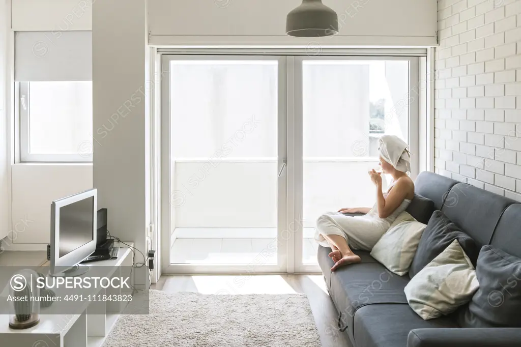 Adult relaxed woman enjoying life while resting on sofa and having hot drink after shower in own cozy apartment