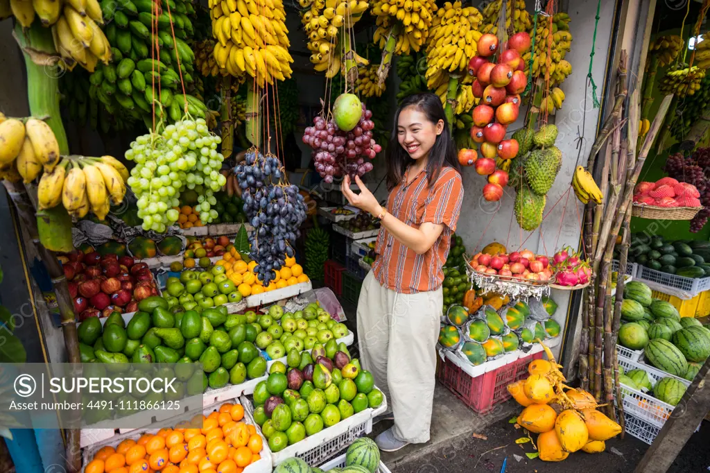 From above of Asian pleased woman on vacation in casual clothing making choice of fresh fruits at market at Sri Lanka;Satisfied Asian resting lady in casual wear tasting grape and smiling at outdoors market