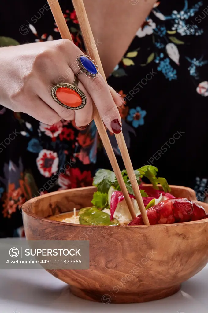 Crop hand with stylish orange and blue rings of faceless woman holding wooden chopsticks while eating Asian noodle bowl;Anonymous fashionable woman with chopstick and Asian dish