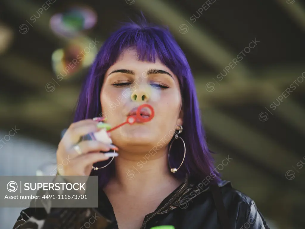 Fashion woman with purple hair smiling and blowing bubbles holding bottle with closed eyes with manicured hands in bright day