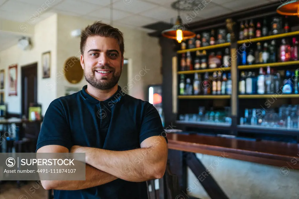 Bright young man in casual wear with crossed arms standing in own bar looking at camera;Happy confident entrepreneur with crossed arms standing in bar