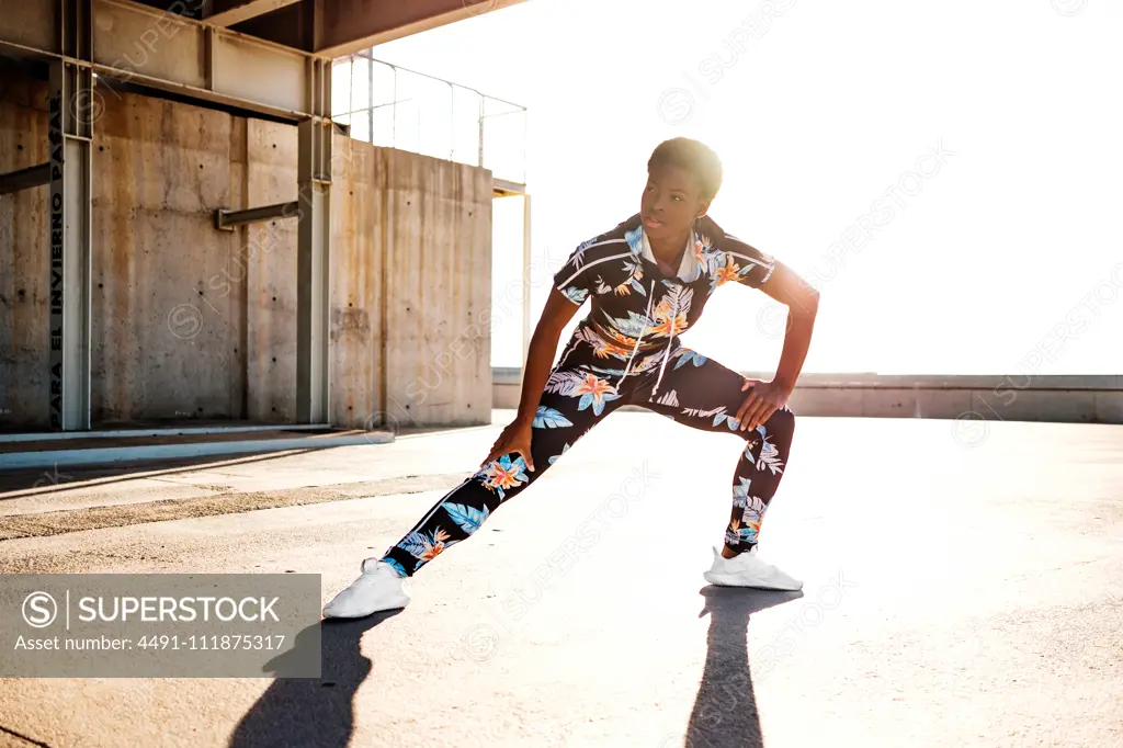 Focused African American adult woman in flowered sportswear stretching leg muscles while standing alone and warming up before training among urban environment in sunny day