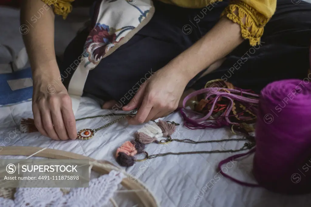 From above of crop female creating handmade dreamcatcher with long threads and accessories sitting in house;Faceless lady creating dreamcatcher at home