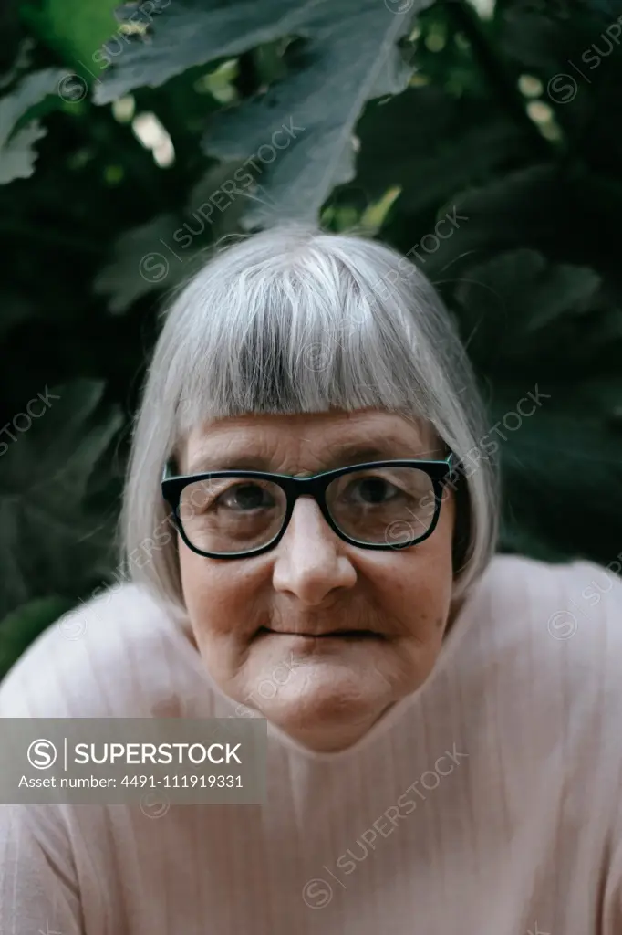 Senior woman with grey hair looking at camera in eyeglasses among plants with big green leafs