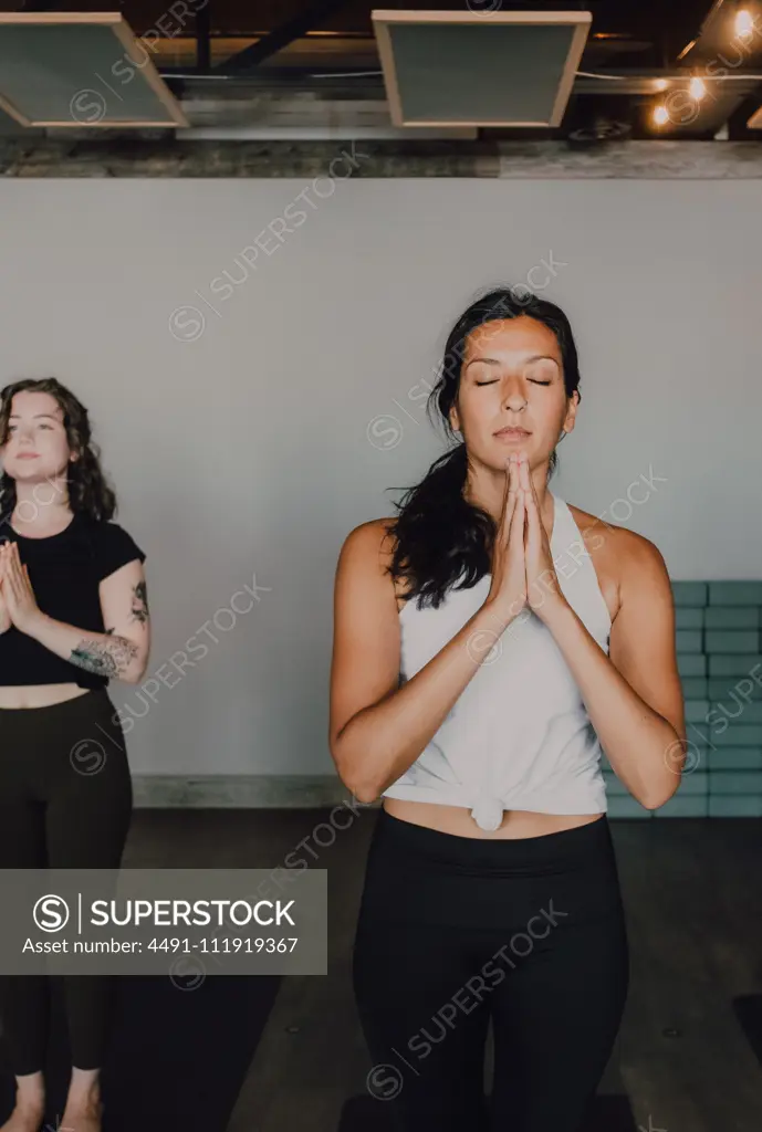Calm slender ladies in activewear with namaste standing in mountain position on sports mats on wooden floor and meditating in spacious contemporary gym with closed eyes