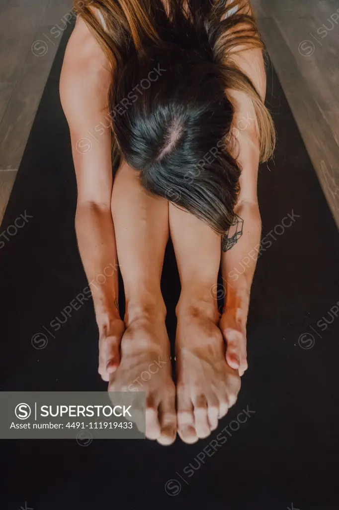 Unrecognizable barefooted female in activewear stretching body while doing paschimottanasana pose sitting on sports mat on floor in modern gym