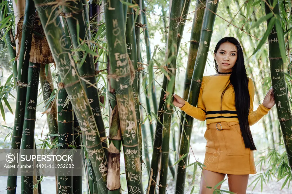 Side view of trendy Asian woman with long dark hair in yellow shirt and short skirt standing in beautiful garden and looking at camera