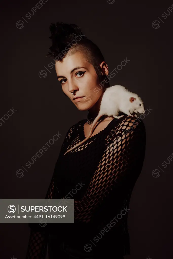 Confident woman with mohawk carrying white rat on shoulder and looking at camera against black background