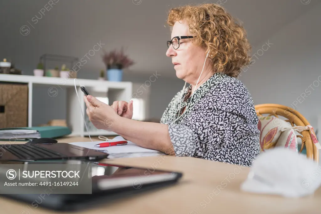 Side view of focused adult woman in casual clothes and eyeglasses using smartphone in earphones while sitting at table and working on laptop at home