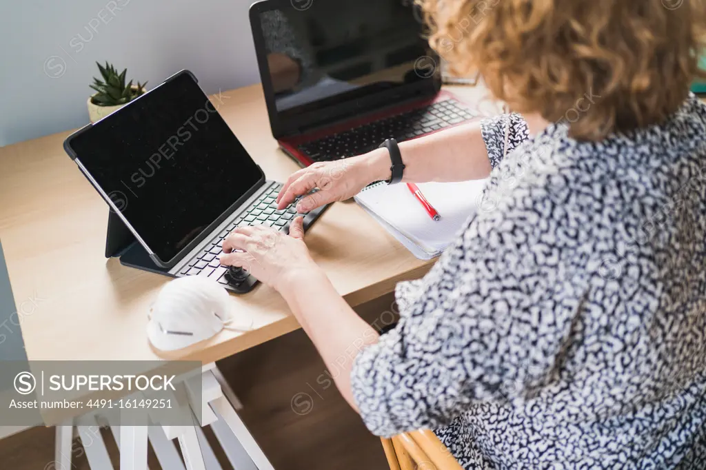 From above view of anonymous female freelancer in casual clothes typing on keyboard while working with laptops at wooden table at home