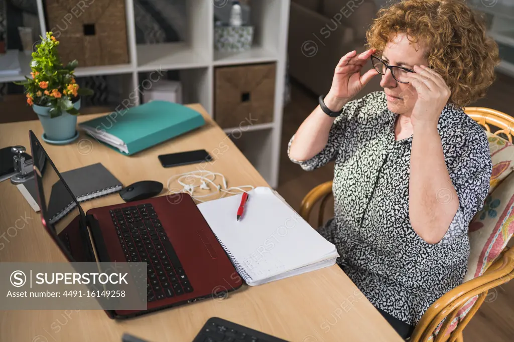 Side view of focused adult woman in casual clothes enjoying music in earphones with closed eyes while resting during work on laptop and notebook