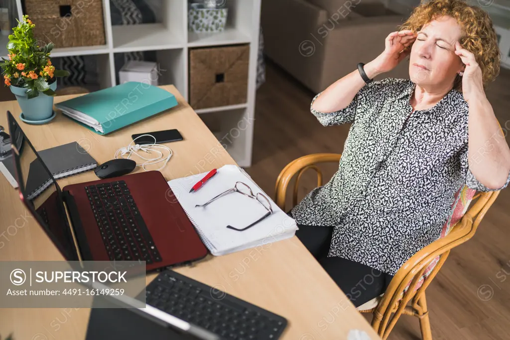 From above view of concentrated adult female freelancer rubbing temples while resting at chair with closed eyes during work with documents and gadgets