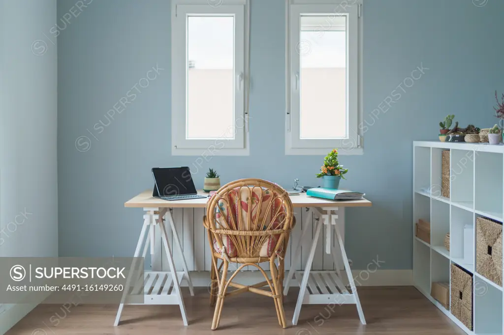 Interior of modern room and workplace with wooden desk with laptop and folder and whicker chair near wooden shelves with boxes and potted plants