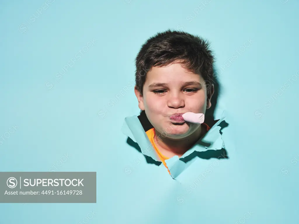 Happy chubby boy with pink candy in mouth smiling and looking at camera through blue ripped paper