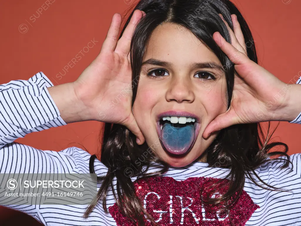 Excited preteen girl in trendy clothes looking at camera and showing blue tongue after eating colored food on red background
